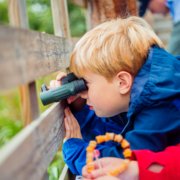 A young child with blonde hair peers through binoculars while leaning against a wooden fence, wearing a blue jacket.